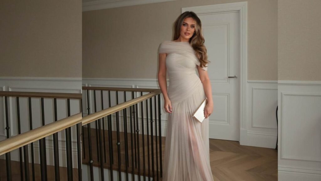 Chloe Lloyd in a flowing long dress stands gracefully on a stairway, showcasing elegance and poise.