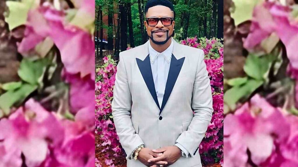 Chris Tucker in a light beige suit with black lapels, jeweled collar, and glasses, stands hands folded before pink blossoms.