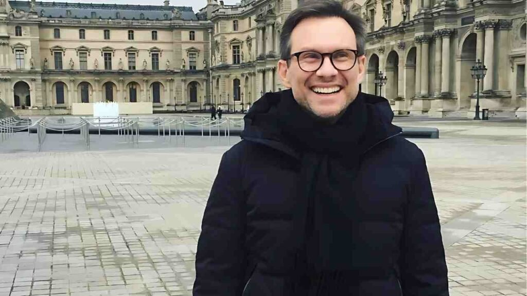 Christian Slater wearing glasses and a black coat stands in front of a large building, looking towards the camera.