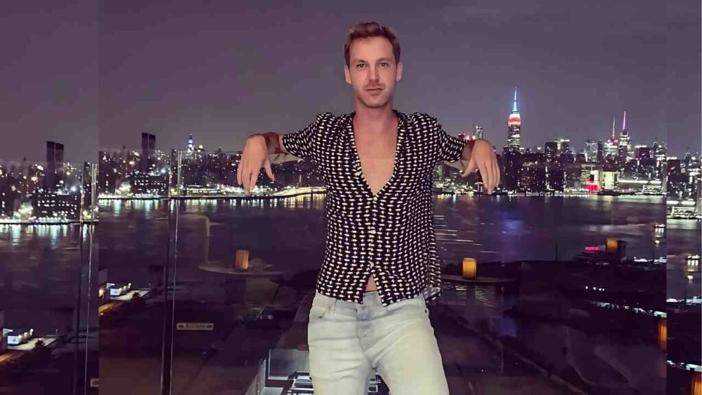 Colin Hanks in a black patterned shirt and light jeans leans on a glass railing at night, city skyline and water behind.