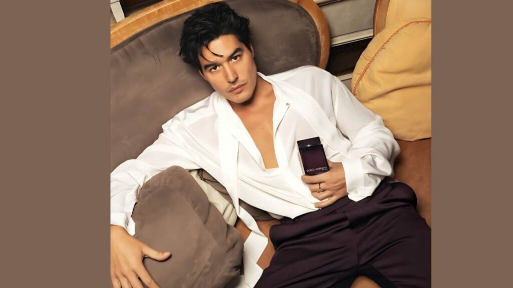 Cristiano Caccamo wearing a white shirt and black pants sits comfortably on a couch, looking relaxed.