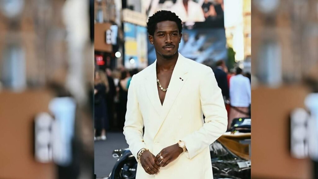 Damson Idris in a white suit stands confidently on a city street, surrounded by urban architecture.