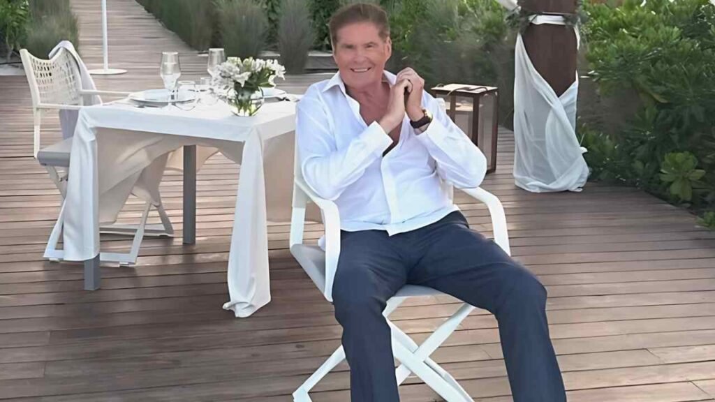 David Hasselhoff in a white shirt and blue pants sits on a chair, looking relaxed and contemplative.