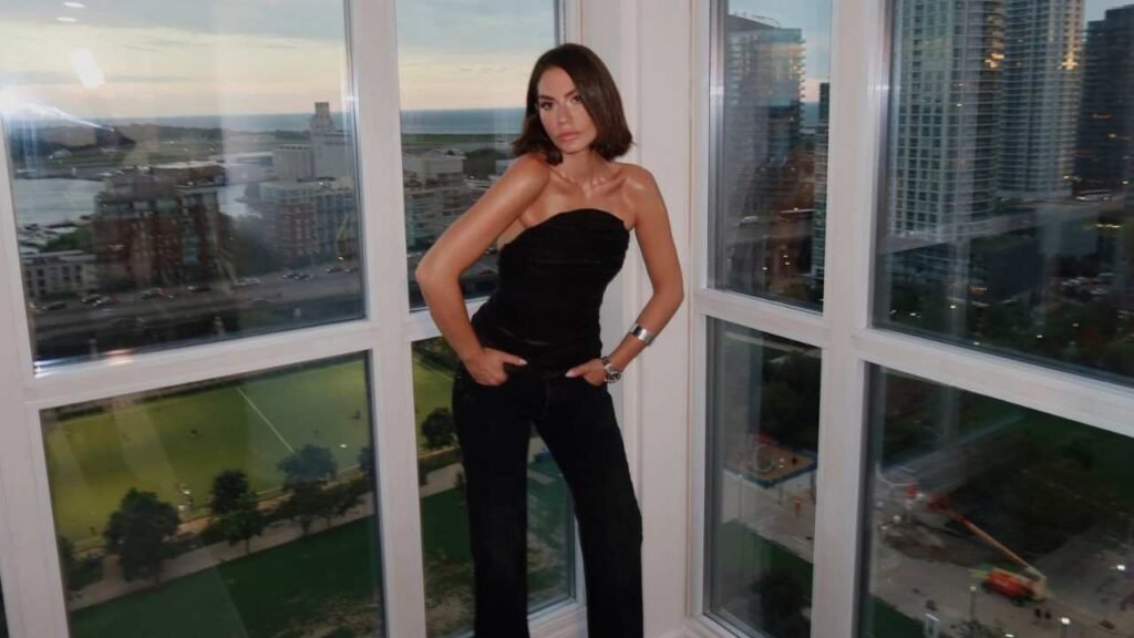 Demet Ozdemir in a black dress stands gracefully in front of a large window, allowing natural light to illuminate the scene.