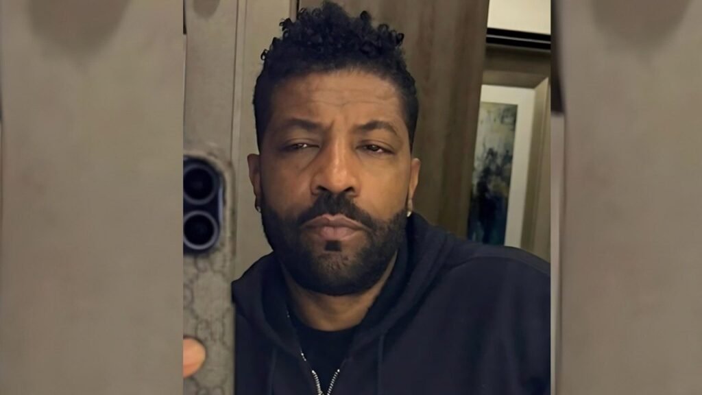 Deon Cole in a black hoodie smiles while taking a selfie with his smartphone.