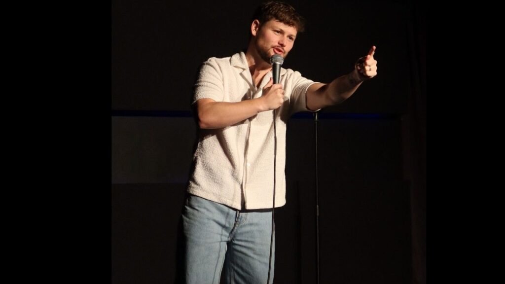 Drew Lynch comedian on stage, holding a microphone and pointing to the audience, wearing a light textured shirt and jeans.