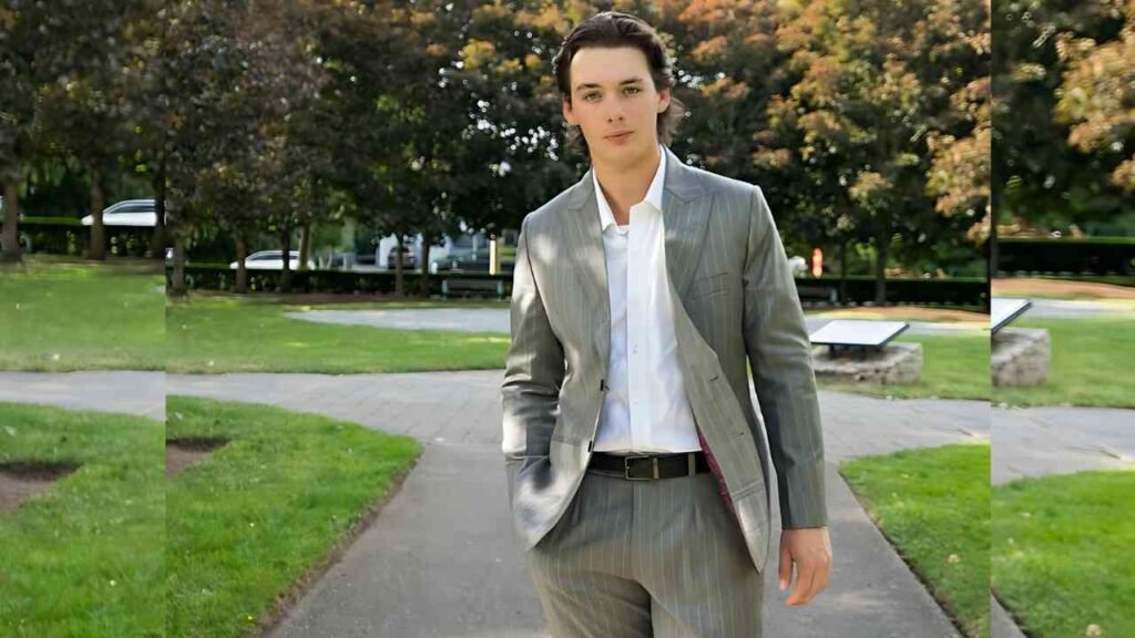 Dylan Kingwell in a suit and tie walking confidently along a paved path surrounded by greenery.