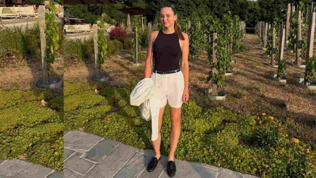 Elena Rybakina wearing a black top and white shorts stands on a stone path surrounded by greenery.