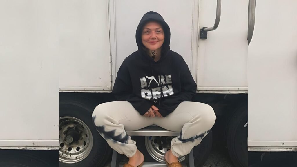 Elle King in a black hoodie with BARE DEN sits on a truck step, wearing tie-dyed pants and tan shoes, before trailer