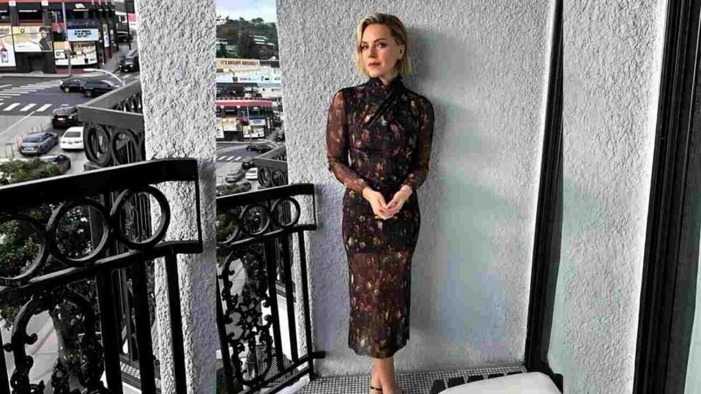 Eloise Mumford in a floral dress stands on a balcony, overlooking a scenic view with greenery in the background.