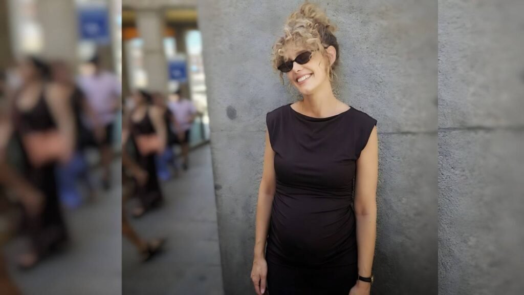Esther Acebo in a black maternity dress leans against a gray wall, sunglasses on, blurred city crowd behind.