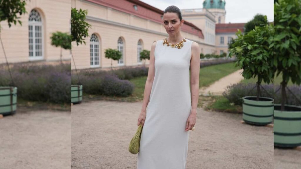 Eva Padberg in a white sleeveless dress with a chunky gold necklace and olive clutch stands in a sunny courtyard.