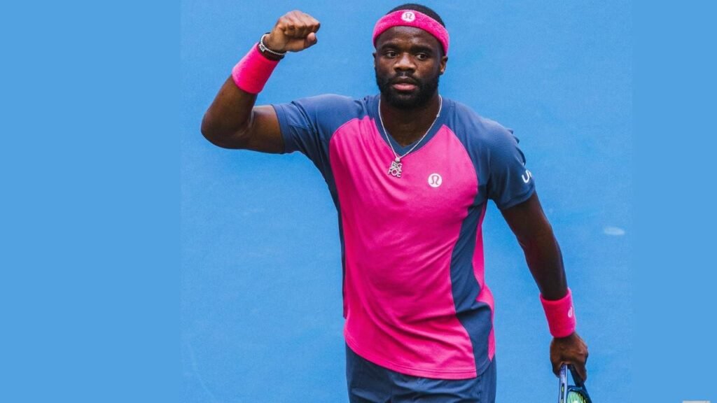 Frances Tiafoe Tennis player in a bright pink and blue outfit raises his left fist in triumph, holding a racket in his right hand against a solid blue backdrop.