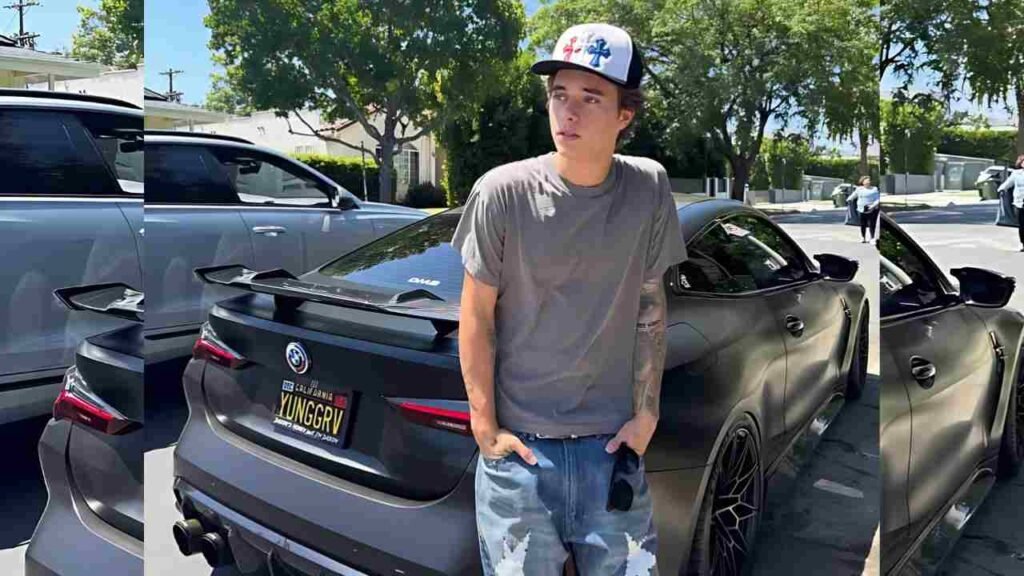 Gavin Magnus in gray tee and ripped denim shorts stands beside matte black BMW with spoiler, plate YUNGGRV, on a sunny street.