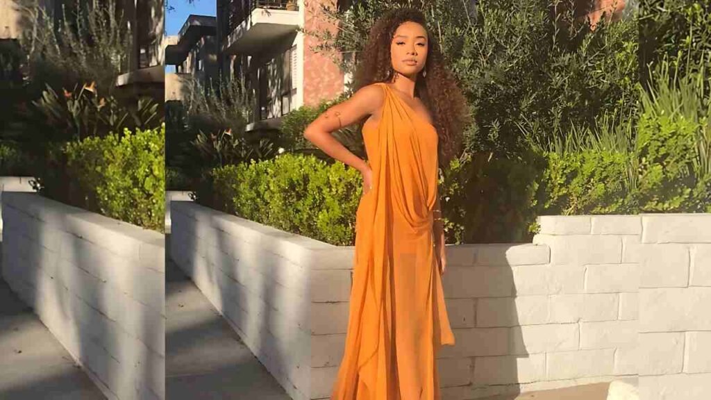 Genneya Walton in an orange draped gown posing by a white brick wall with sunlit greenery behind.