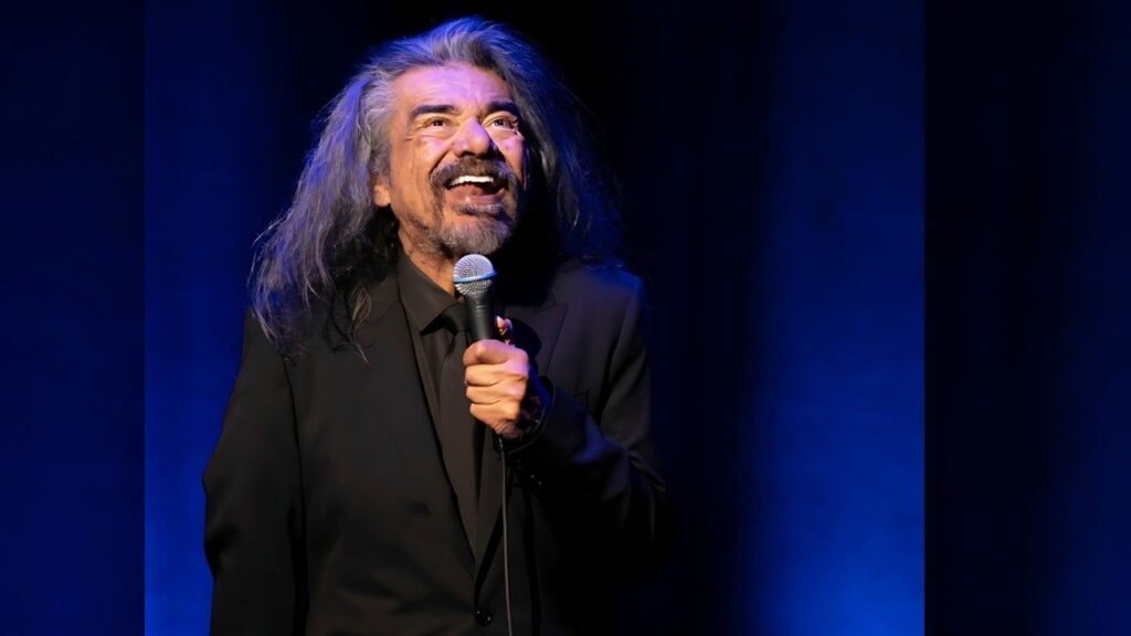 George Lopez with long hair stands with a microphone, indicating he is about to address an audience.