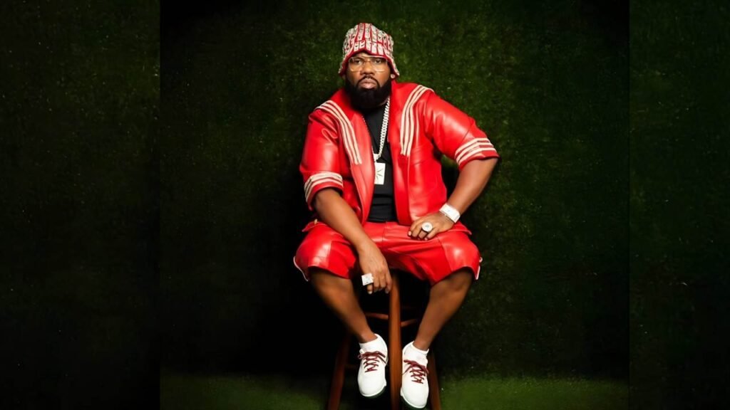 Ghostface Killah in a red shiny jacket and shorts sits on a wooden stool, wearing a patterned bucket hat, against a dark green backdrop.