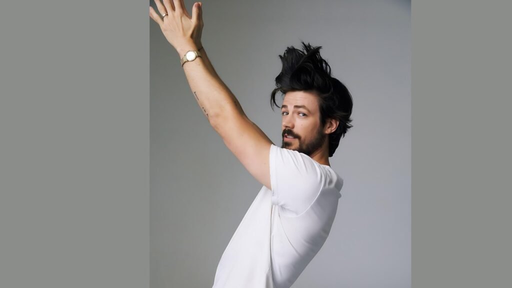 Grant Gustin in a white shirt is reaching upward, possibly to grab something from a high shelf.