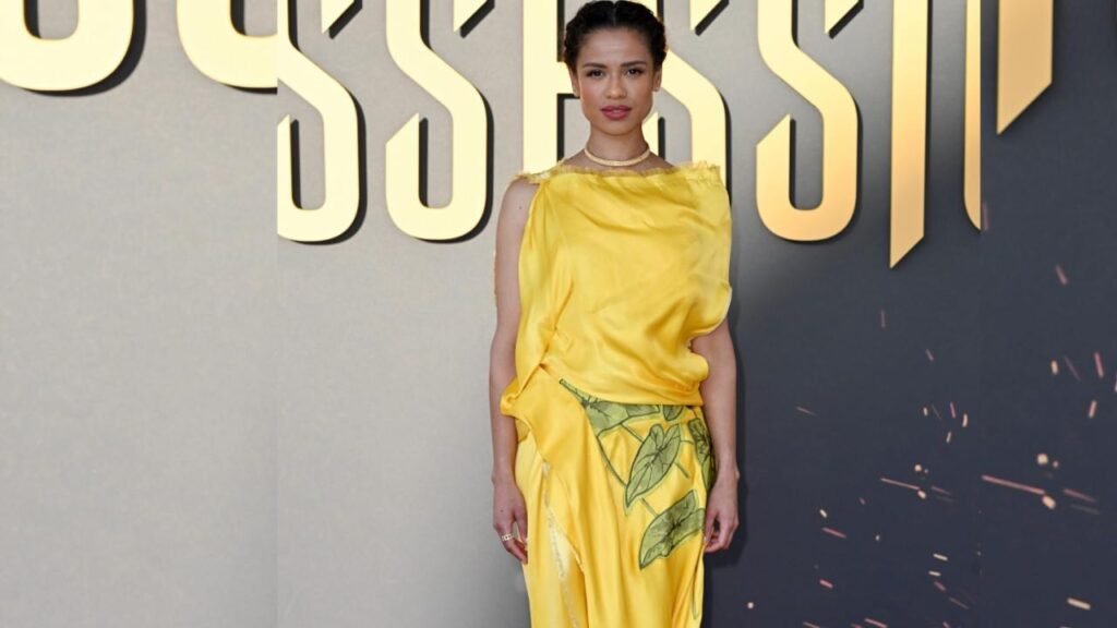 Gugu Mbatha Raw in a yellow satin top and leaf-print skirt poses before a backdrop of large gold lettering.