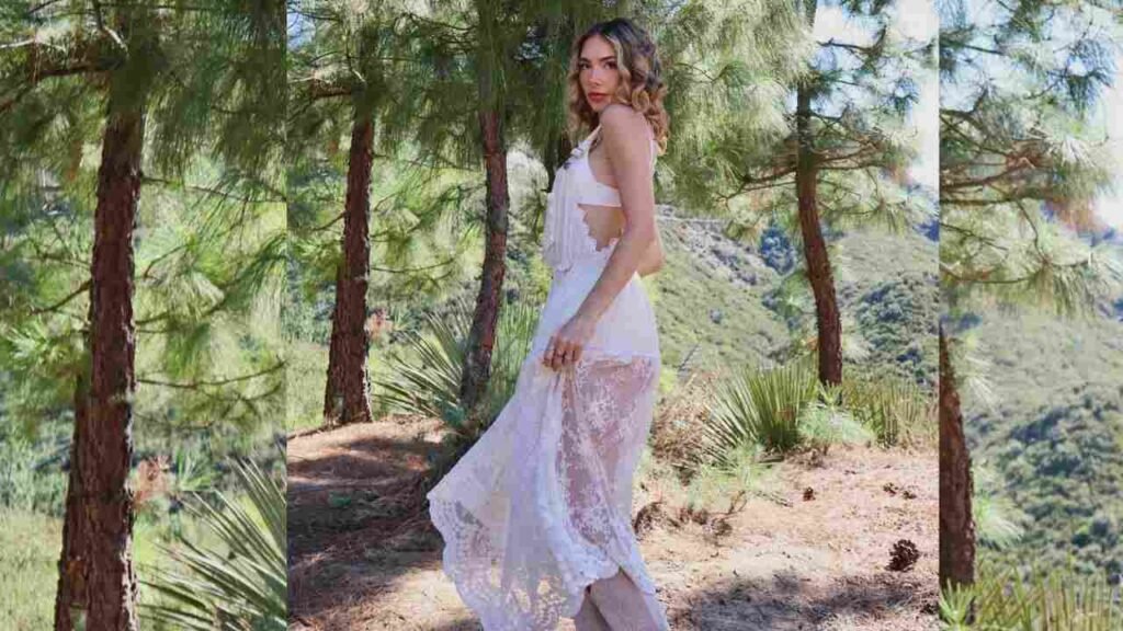 Haley Pullos in a flowing white dress stands gracefully among tall trees in a serene forest setting.