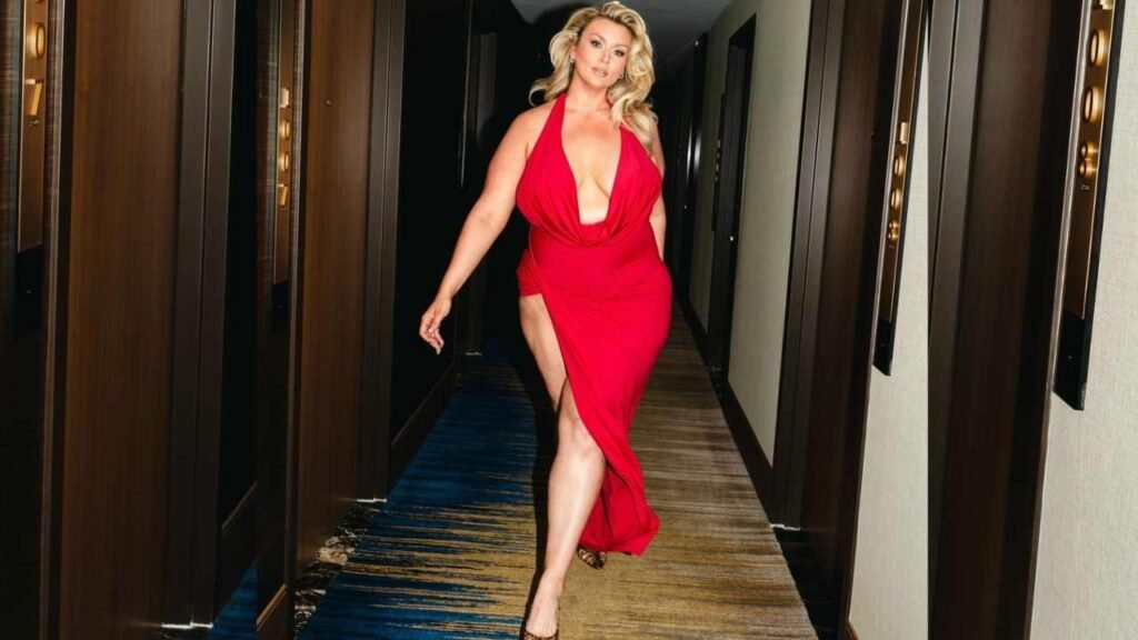 Hunter McGrady in a red dress walks gracefully down a well-lit hallway.