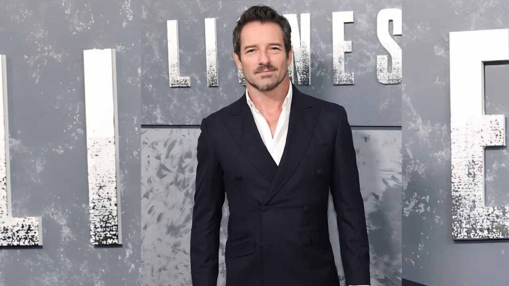 Ian Bohen in a formal suit and tie stands confidently on a vibrant red carpet, showcasing elegance and sophistication.