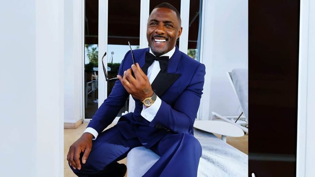 Idris Elba in a blue suit and tie is seated on a chair, looking confidently at the camera.