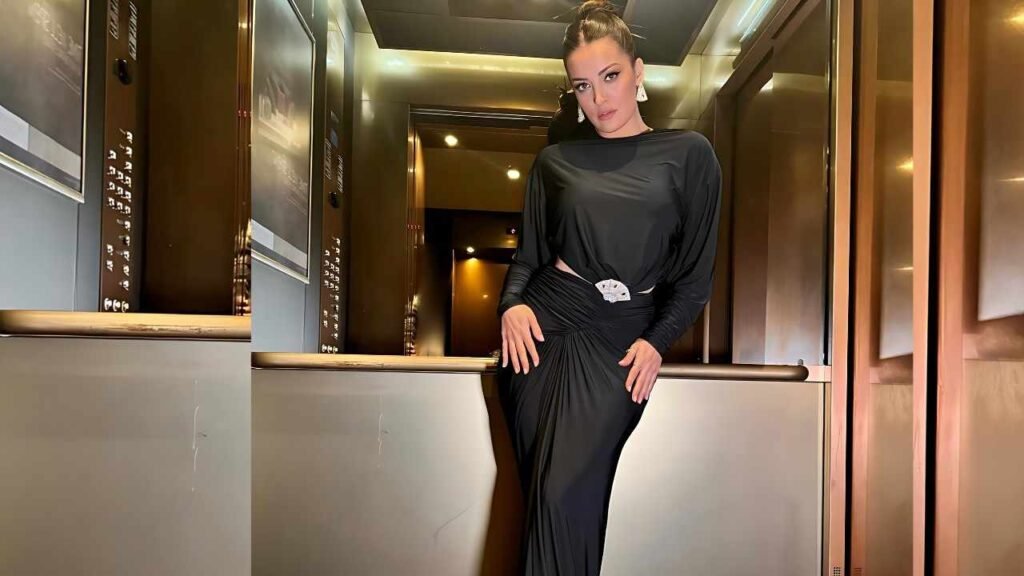 Irem Sak in a black long-sleeve top and ruched high-waisted skirt leans on a metal elevator railing, warm lights glow.