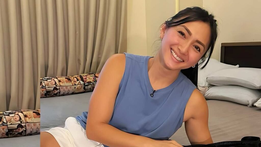 Iya Villania in blue sleeveless top and white shorts lounges on bed, lapel mic clipped to her top, beige curtains behind.