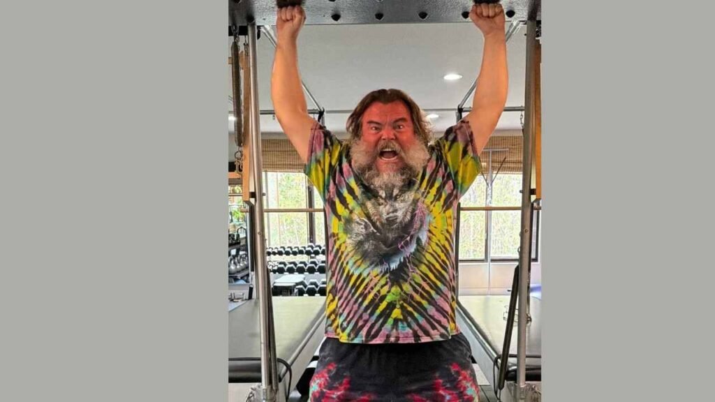 Jack Black in a tie-dye shirt performs a pull-up on a gym bar, showcasing strength and determination.