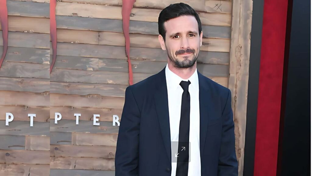 James Ransone in a dark navy suit and black tie smiles at a wooden paneled backdrop with red ribbons.