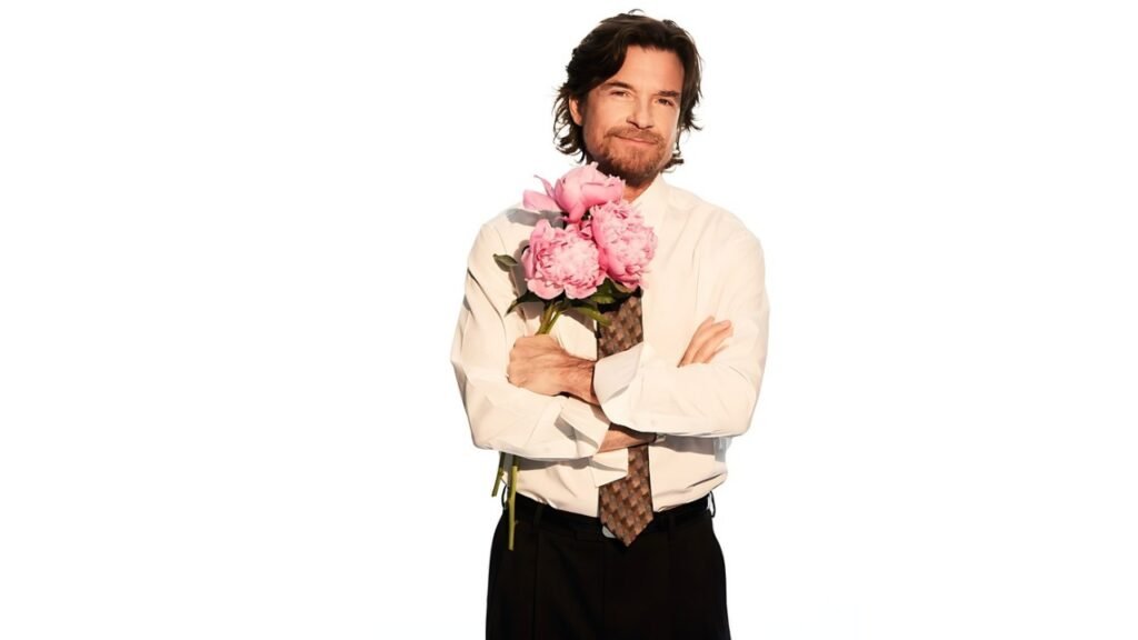 Jason Bateman in a shirt and tie holds a colorful bouquet of flowers, smiling warmly at the camera.