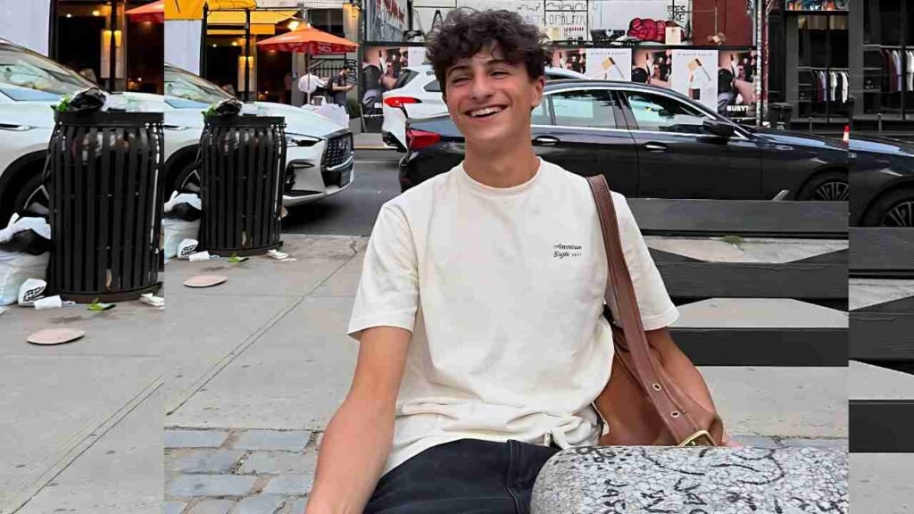 Judd Goodstein in a cream T-shirt and dark jeans sits on a bench by a busy city sidewalk, brown bag over his shoulder.