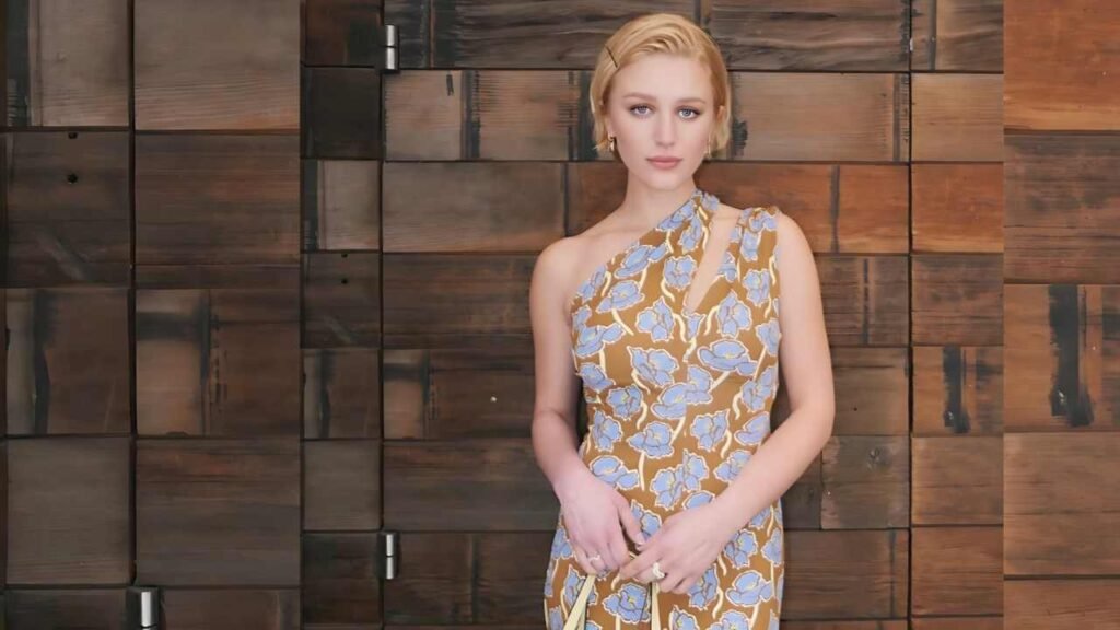Julia Schlaepfer in a mustard one-shoulder dress with blue floral pattern stands before a wooden wall, hands clasped.