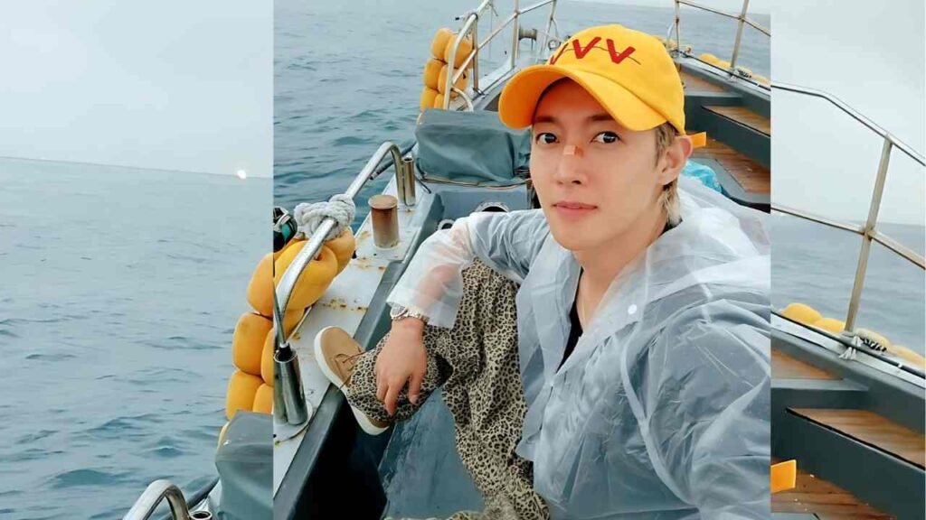 Kim Hyun-joong in poncho, leopard pants, and a yellow cap, with yellow life jackets on rail and sea behind.