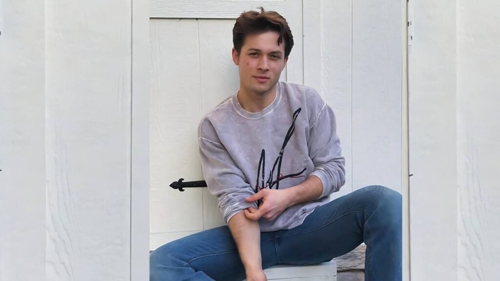 Leo Howard sits on a bench, hands resting on his knees, looking contemplative in a park setting.