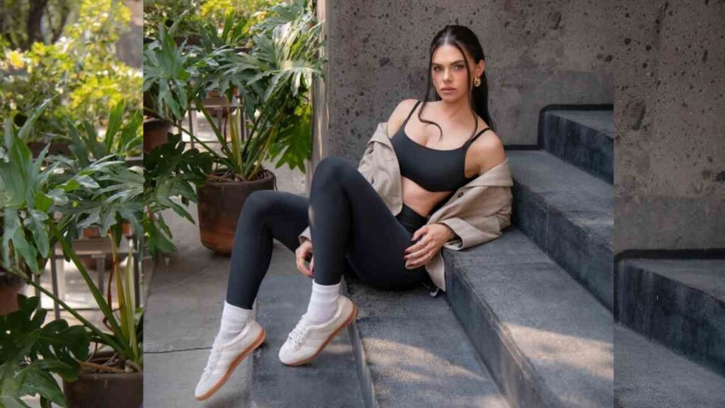 Livia Brito sits on gray stone steps in a black sports bra and leggings, with a beige jacket and white sneakers; plants around.