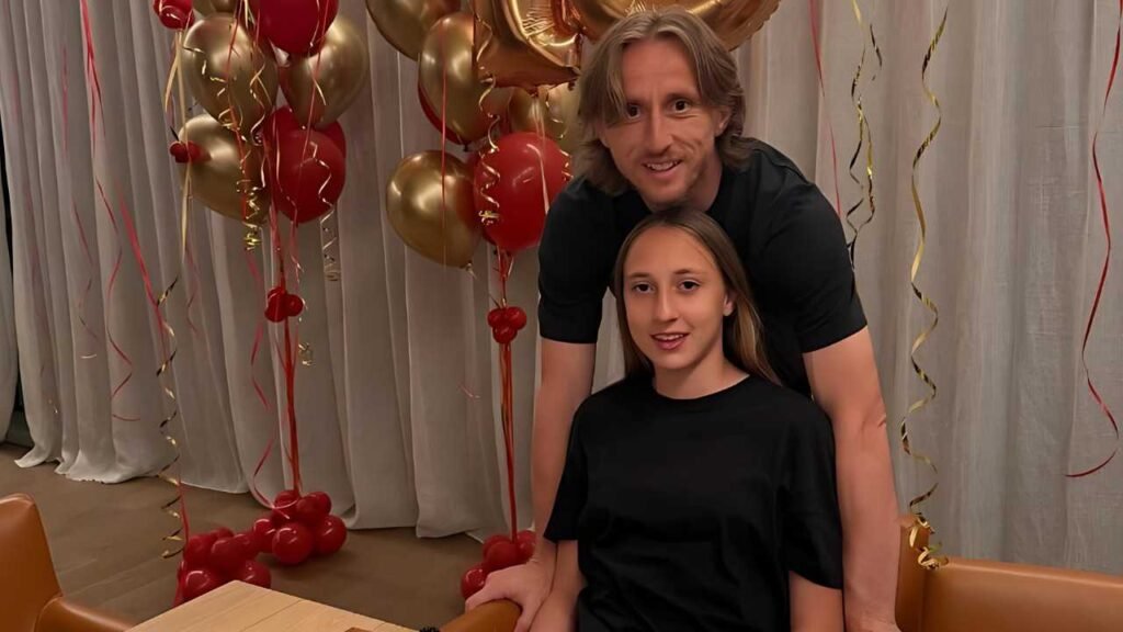 Luka Modric leans over a seated young woman at a table, surrounded by red and gold balloons and streamers in a festive room.