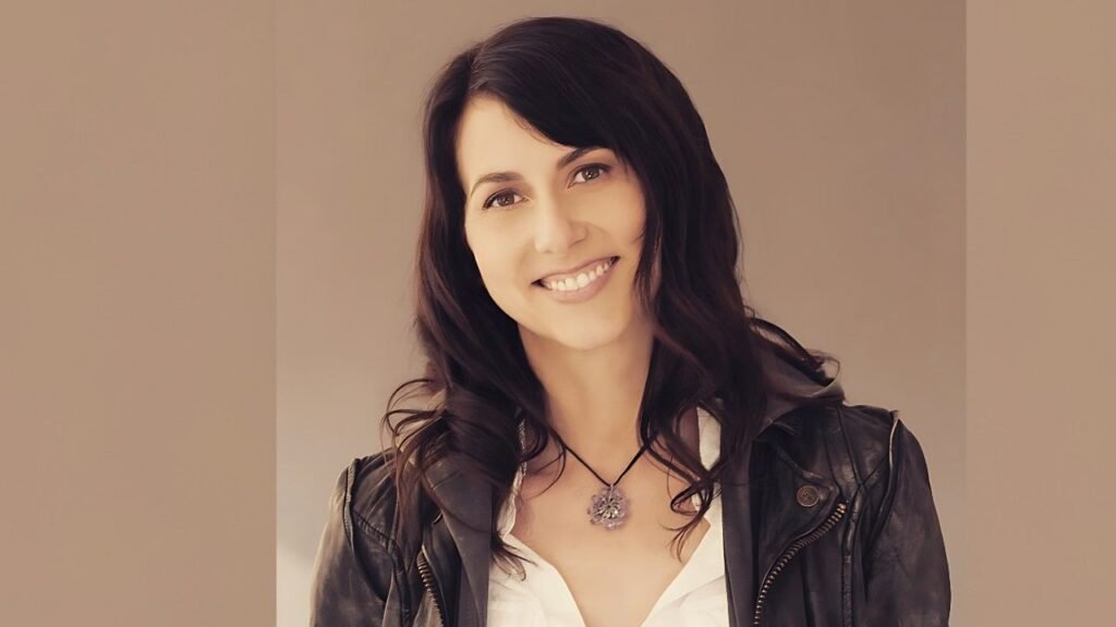 Mackenzie Bezos with dark hair wearing a black leather jacket over a white blouse and pendant necklace against beige backdrop.