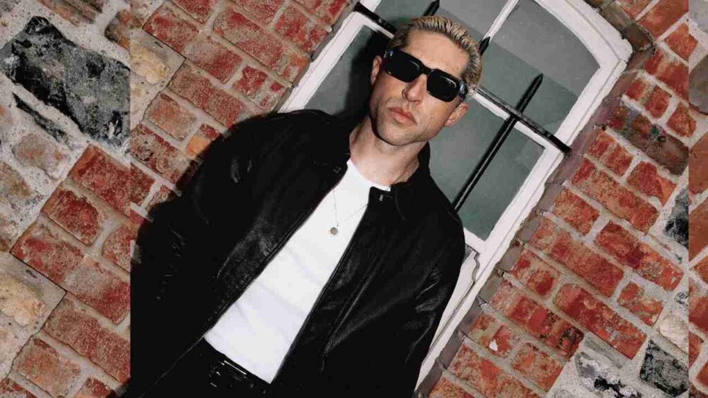 Michael Shuman wearing sunglasses stands beside a textured brick wall, showcasing a casual and confident demeanor.