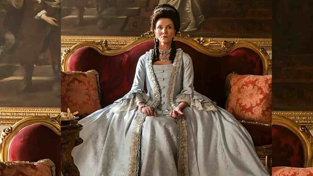 Michelle Fairley in a blue 18th-century gown sits on a gilded red velvet throne, hands folded, ornate jewelry, in an opulent room.