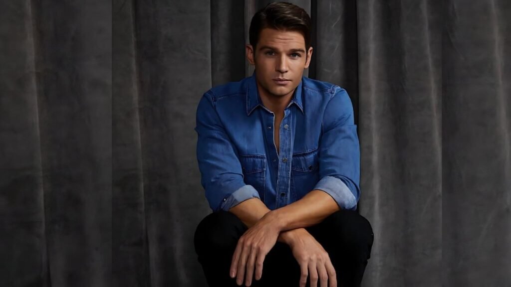 Mike Vogel seated cross-legged in a blue denim shirt, hands resting on his knees, against dark gray curtains.