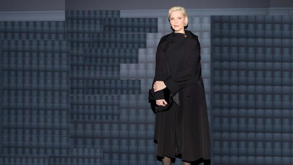 Nadja Auermann with short platinum hair in a long black coat, clutch in hand, stands before a blue-gray grid of square panels.
