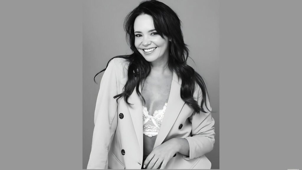 Natasha Klauss with long dark hair wearing a light, double-breasted blazer over lace top, posed against a monochrome backdrop.