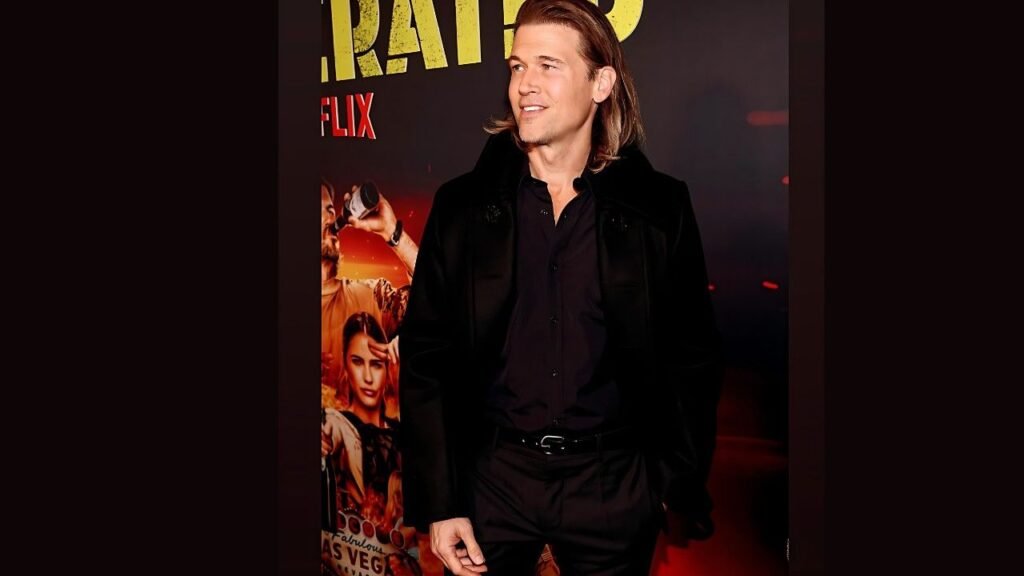 Nick Zano with shoulder-length blond hair in a black shirt and velvet jacket stands on a red carpet, smiling before a warm poster.