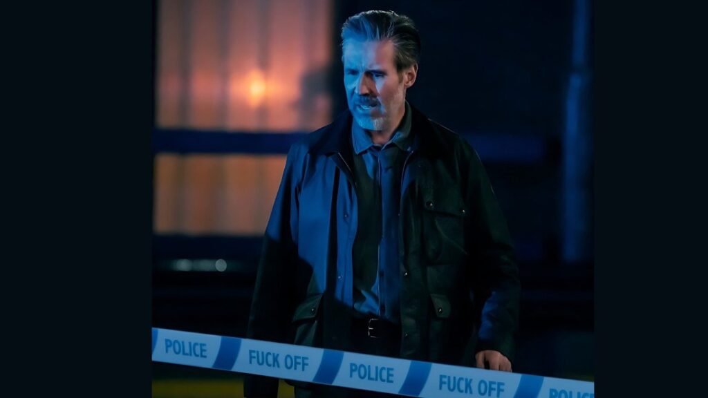Paddy Considine in a jacket stands in front of police tape, indicating a potential crime scene or investigation area.