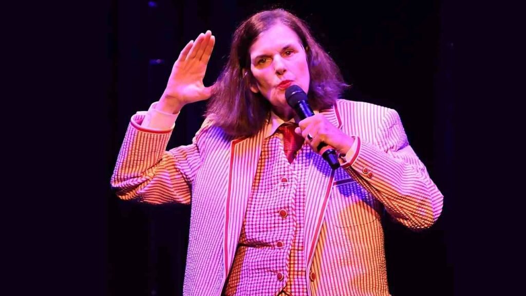 Paula Poundstone in a suit and tie stands confidently, holding a microphone, ready to speak or present.