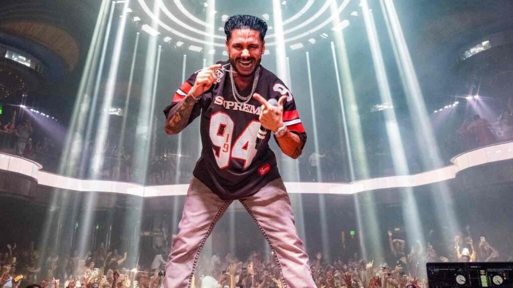 Pauly D Performer on an arena stage, wearing a black Supreme jersey with 94, flashing a medal as lasers cut through a cheering crowd.