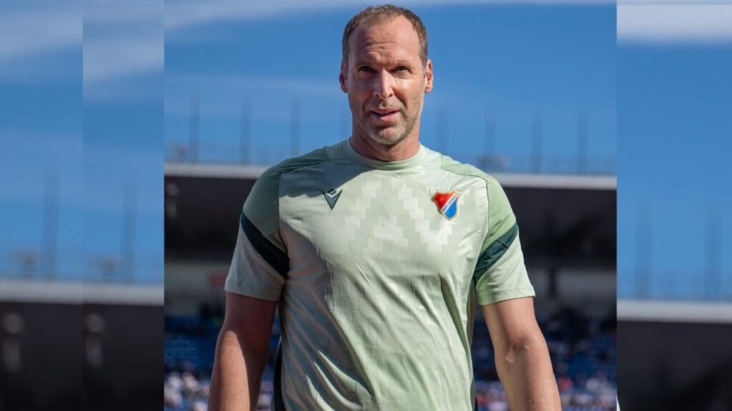 Petr Cech wearing a green shirt stands on a soccer field, surrounded by grass and goalposts in the background.
