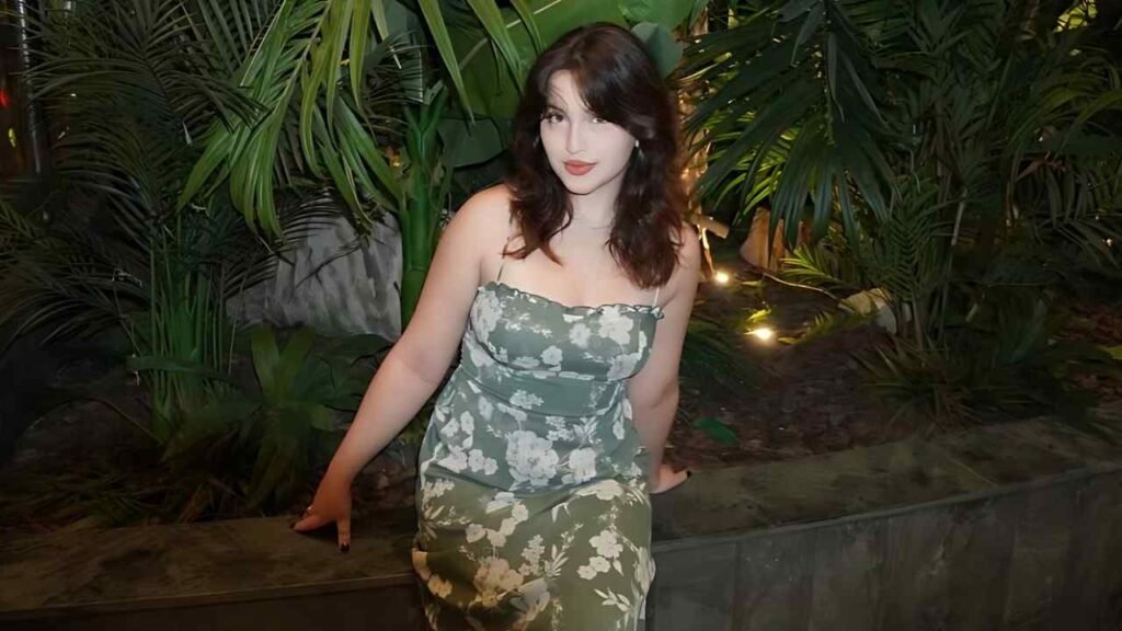 Righley Engelhardt in a green floral strapless dress sits on a low wall among lush tropical plants, softly lit by night garden lights.